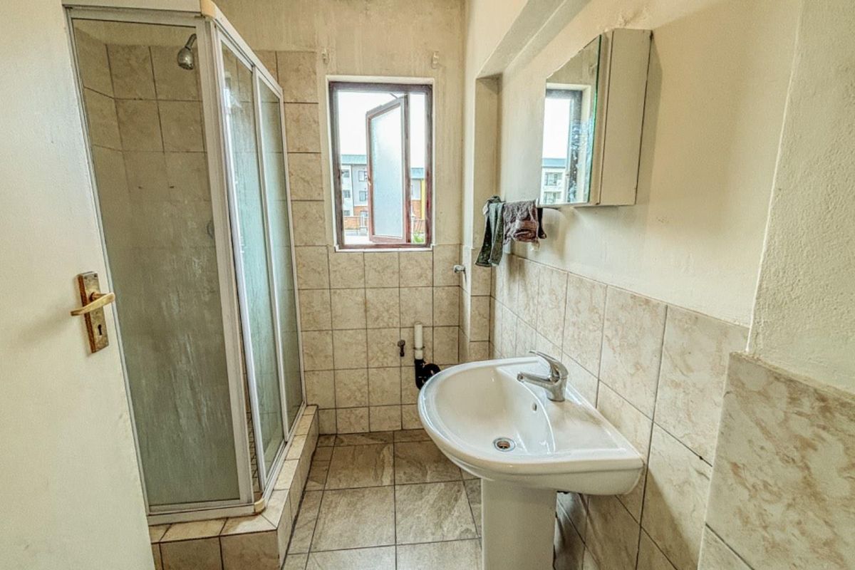 A compact bathroom featuring a pedestal sink, a corner shower enclosure with tiled surround, and a small window for natural ventilation.