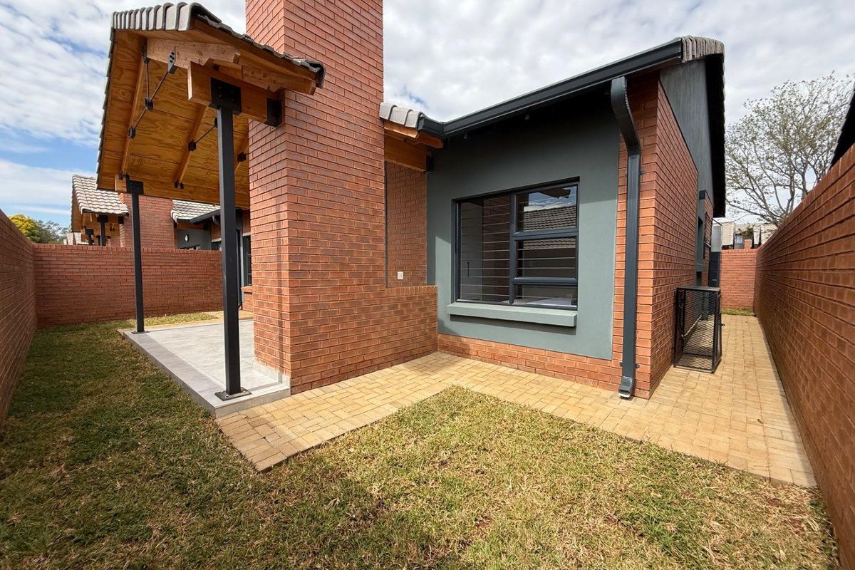A well-maintained rear exterior view of the townhouse featuring a covered patio, brick chimney, paved walkways, and a private garden area.