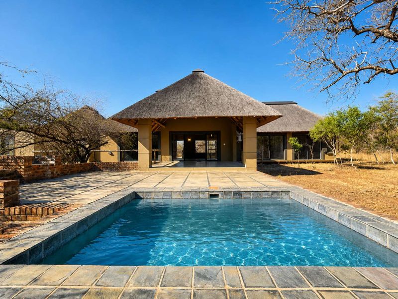 Rear view of a thatched-roof house featuring a swimming pool, paved patio area, and sliding glass doors leading into the main living space.