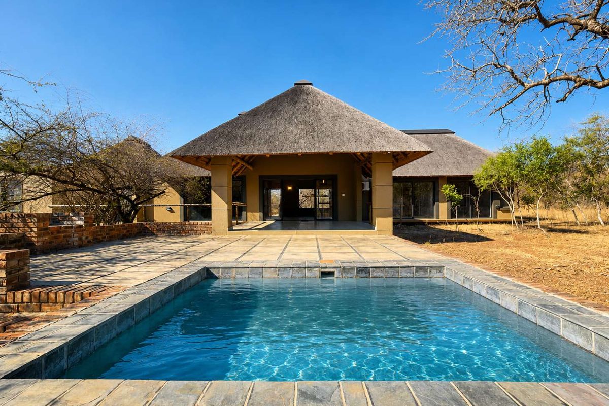 Rear view of a thatched-roof house featuring a swimming pool, paved patio area, and sliding glass doors leading into the main living space.