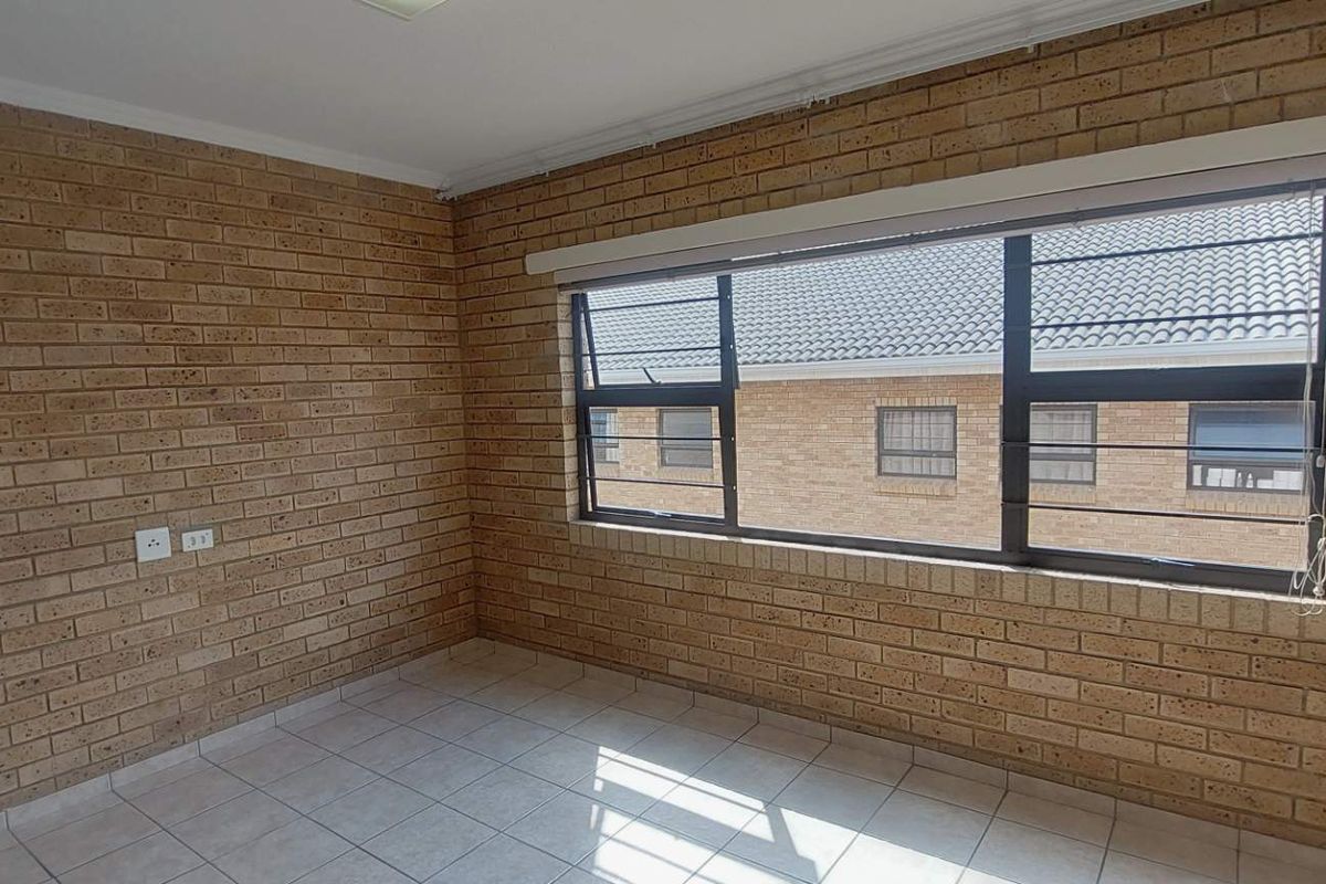 A bright, empty bedroom featuring exposed brick walls, tiled flooring, and large windows with security bars and a roller blind.
