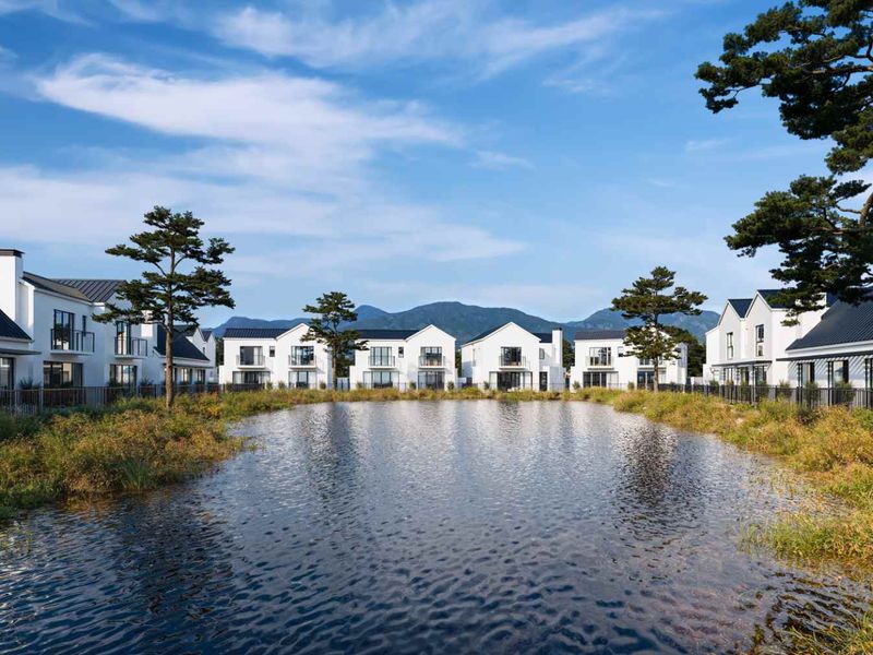 A scenic view of a modern residential estate featuring white houses with metal roofs, balconies, and a central pond surrounded by landscaped greenery.