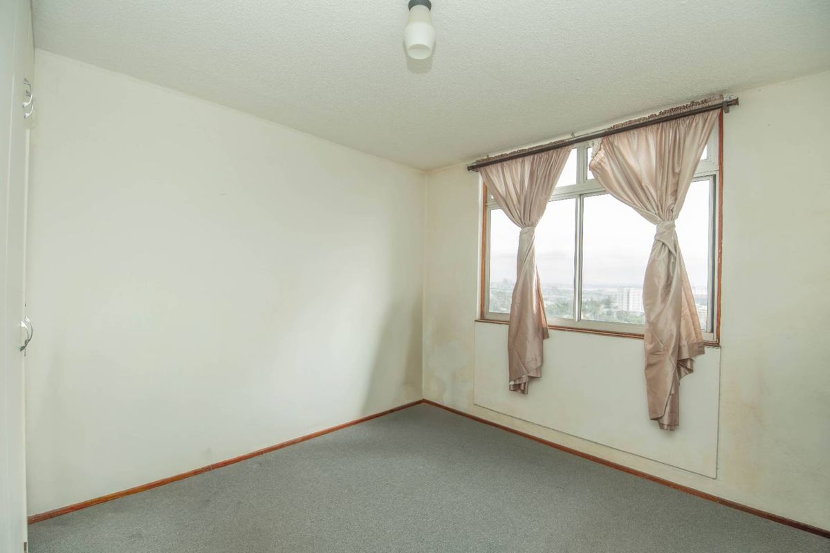 An empty bedroom featuring carpeted flooring, a built-in cupboard, and a window with views, showing signs of wall staining and wear.