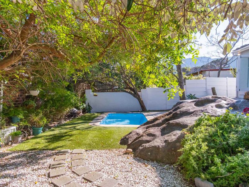 A private backyard featuring a swimming pool, artificial lawn, and natural rock landscaping, enclosed by a white boundary wall.