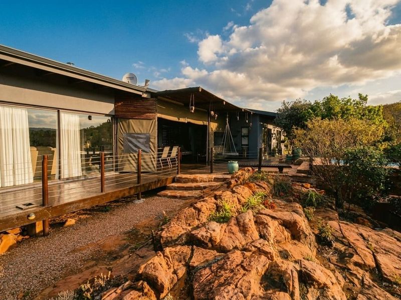 A spacious rear exterior featuring a wooden deck, covered patio area, and natural stone landscaping, offering a seamless transition between indoor and outdoor living spaces.