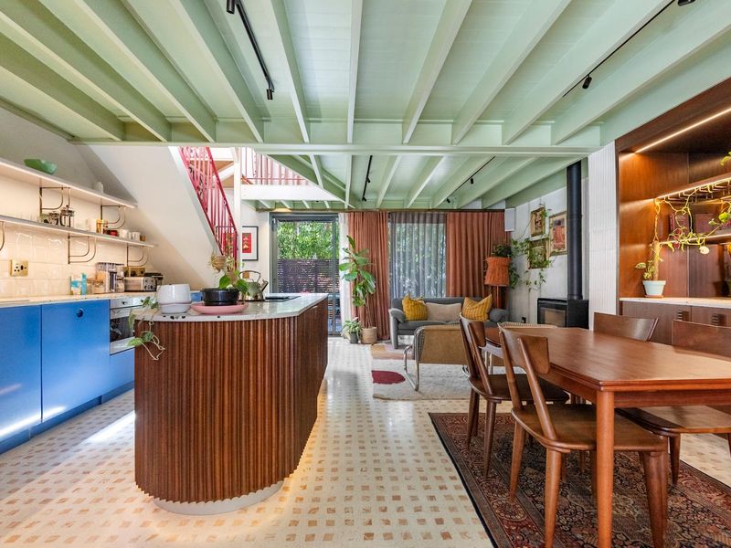 A stylish open-plan living and kitchen area featuring a fluted timber island, exposed green beams, a fireplace, and elegant terrazzo flooring.