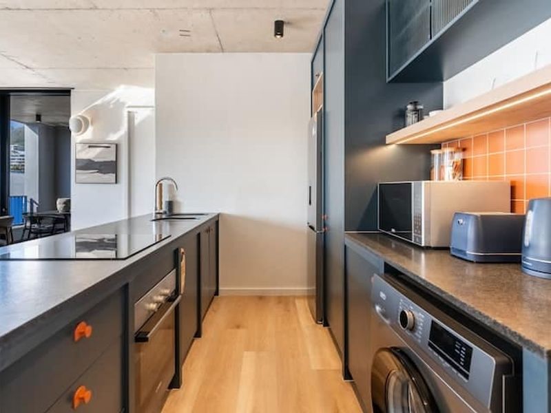 Modern kitchen featuring dark cabinetry, stone countertops, integrated appliances, and a vibrant tiled backsplash, set against an exposed concrete ceiling.