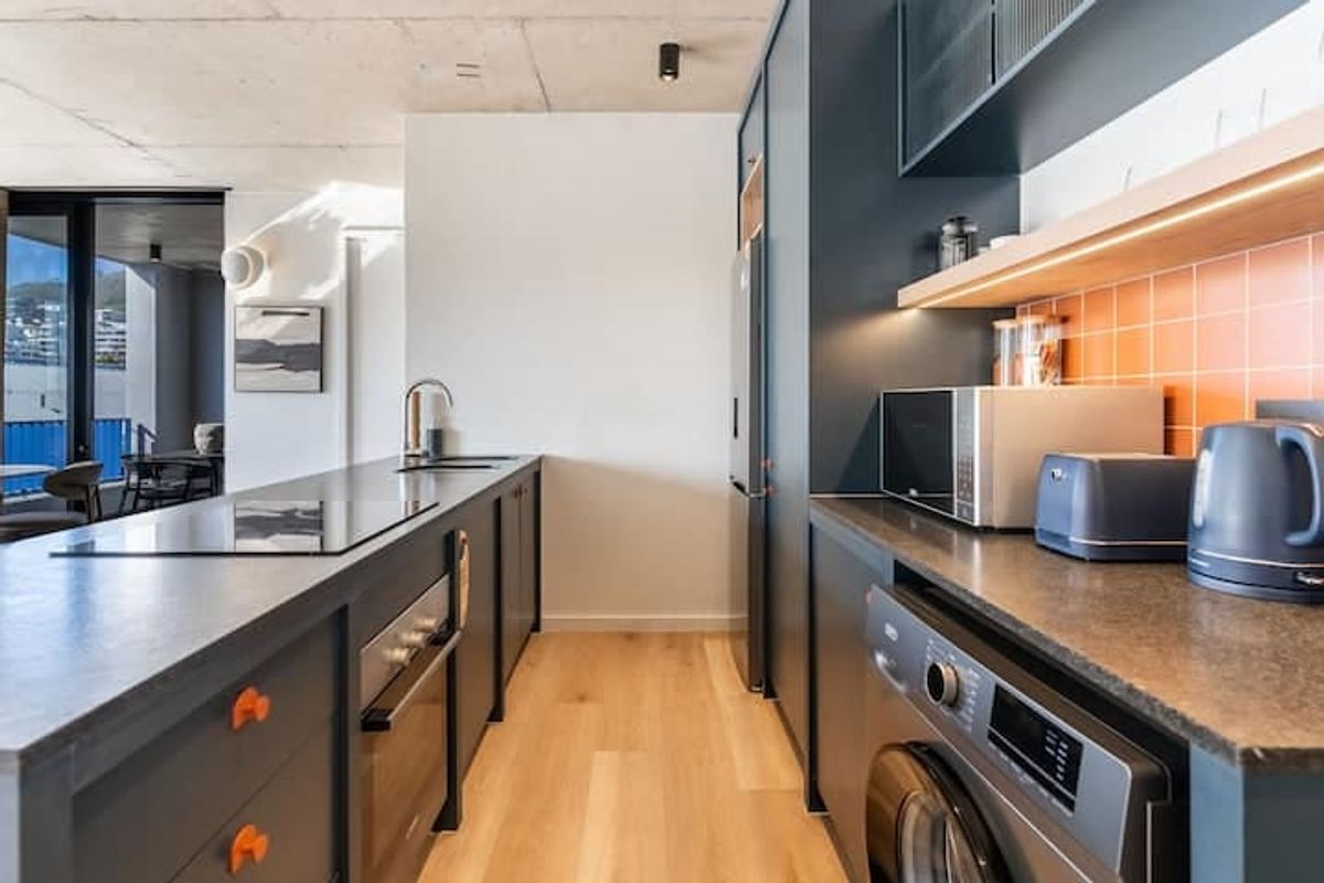 Modern kitchen featuring dark cabinetry, stone countertops, integrated appliances, and a vibrant tiled backsplash, set against an exposed concrete ceiling.