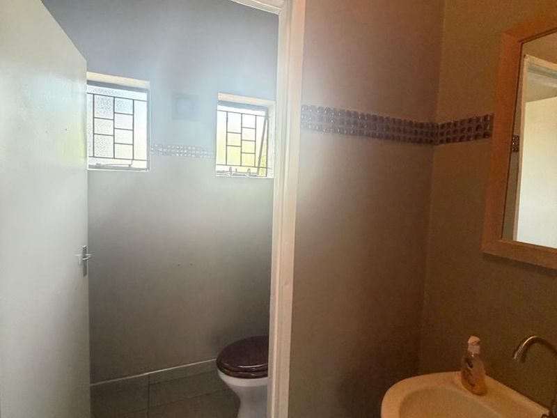 A functional bathroom featuring a washbasin with a wall-mounted mirror and a separate toilet area with tiled flooring and security-barred windows.