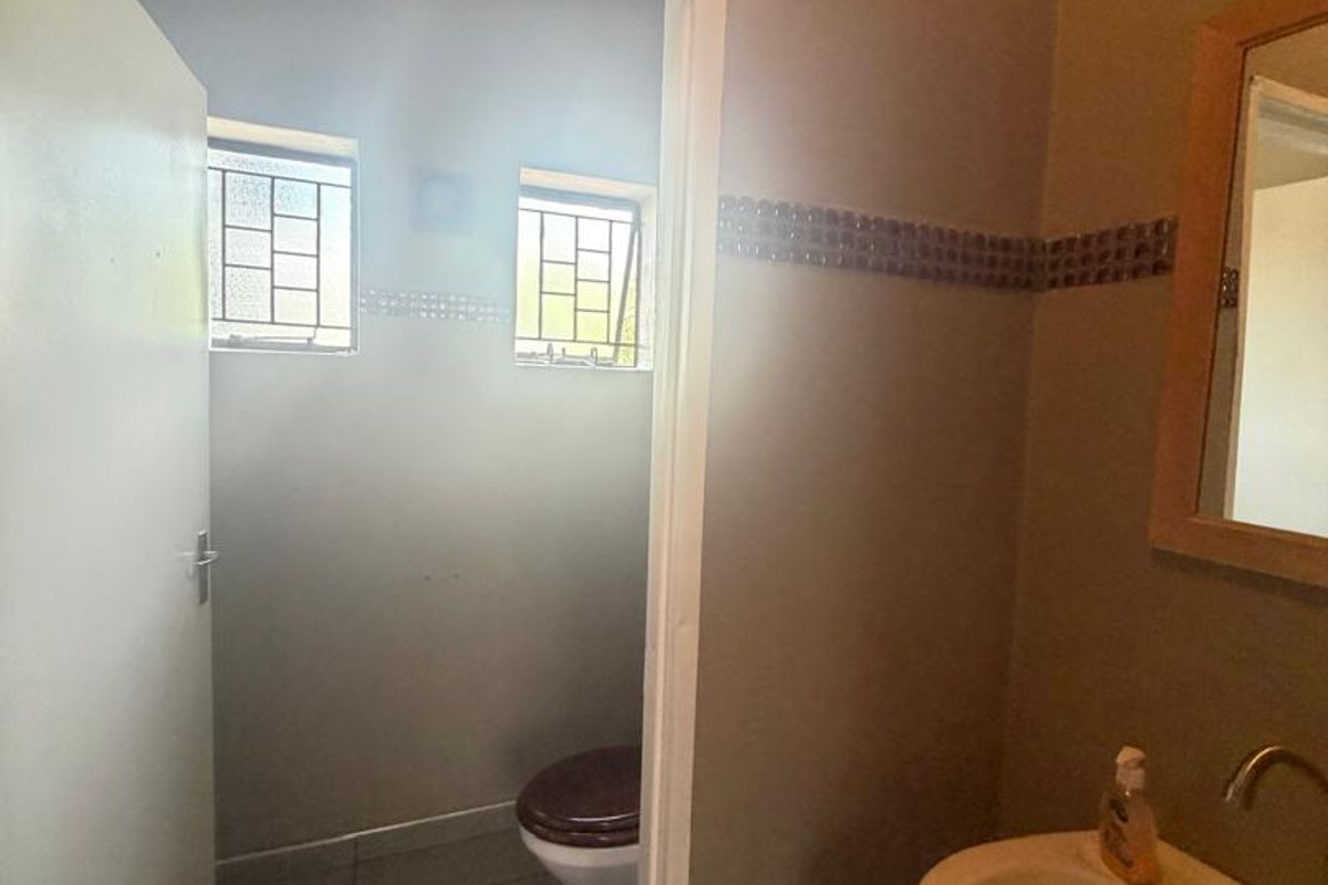 A functional bathroom featuring a washbasin with a wall-mounted mirror and a separate toilet area with tiled flooring and security-barred windows.