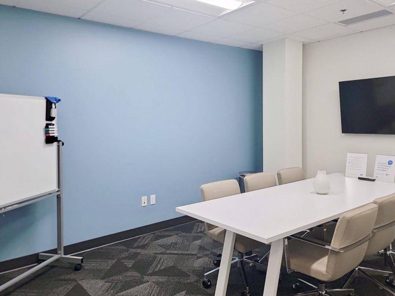 A professional office meeting room featuring a wall-mounted television, carpeted flooring, and a suspended ceiling with recessed lighting.