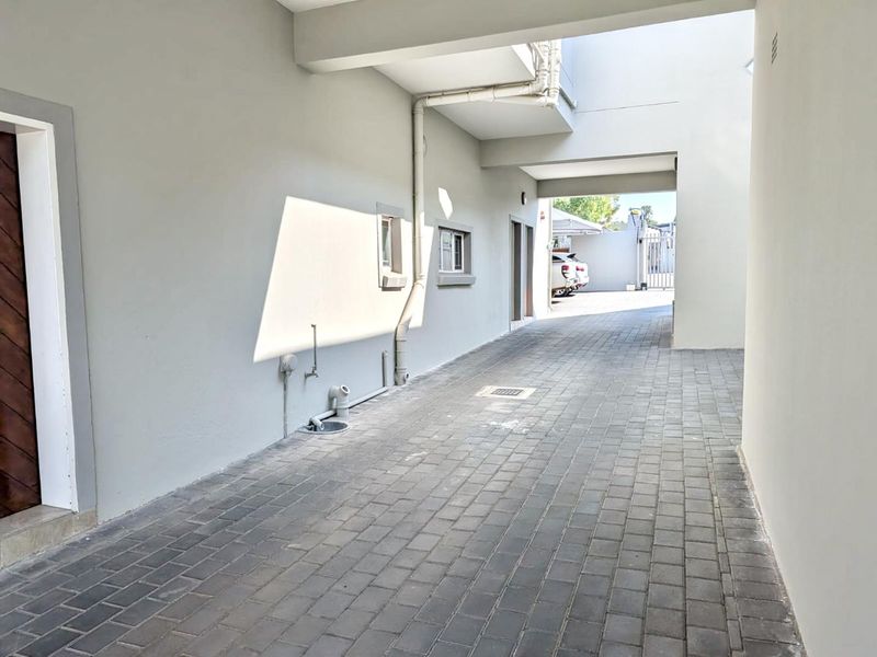 A clean, paved exterior walkway or driveway area featuring neutral walls, external plumbing fixtures, and a covered section leading to the rear of the property.