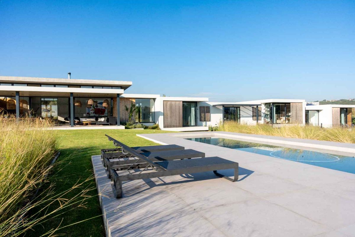 Modern architectural rear elevation featuring a swimming pool, expansive paved patio, and seamless indoor-outdoor flow through large glass sliding doors.