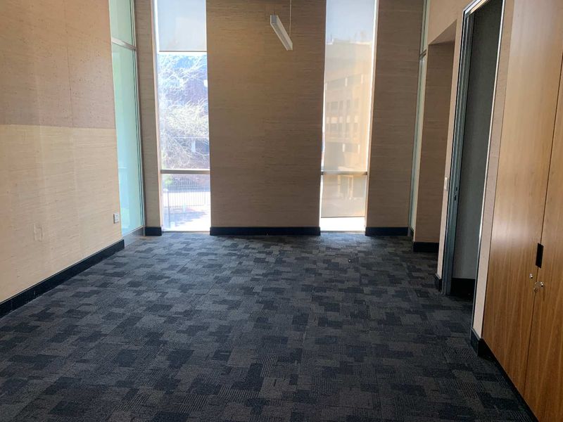 A bright, empty office space featuring floor-to-ceiling windows, patterned carpet flooring, and built-in wooden cabinetry.