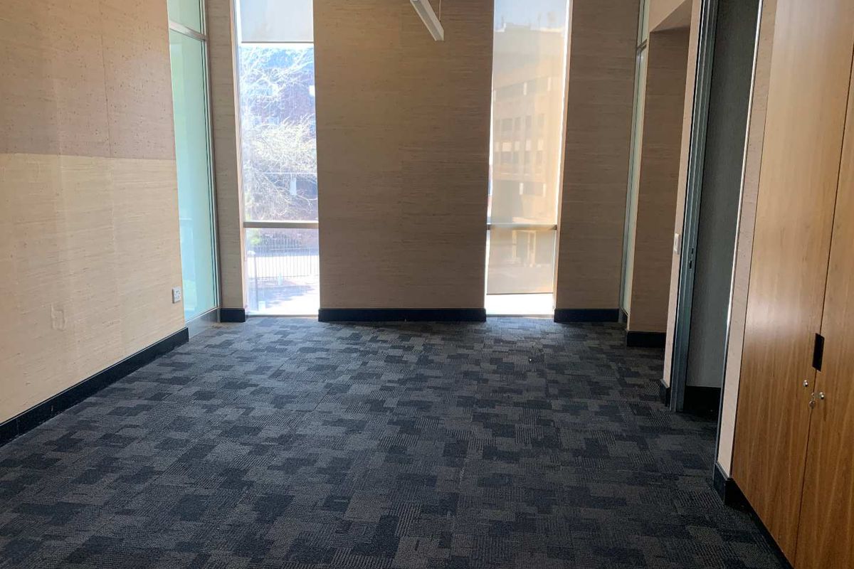A bright, empty office space featuring floor-to-ceiling windows, patterned carpet flooring, and built-in wooden cabinetry.