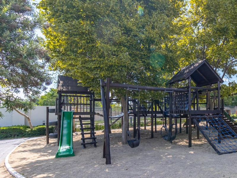 A well-maintained communal garden area featuring a wooden playground structure, sand pit, and paved pathways surrounded by mature trees.