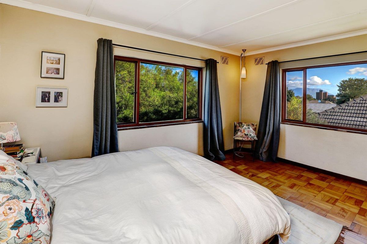 A well-lit bedroom featuring classic parquet flooring, large windows with natural light, and neutral wall finishes.