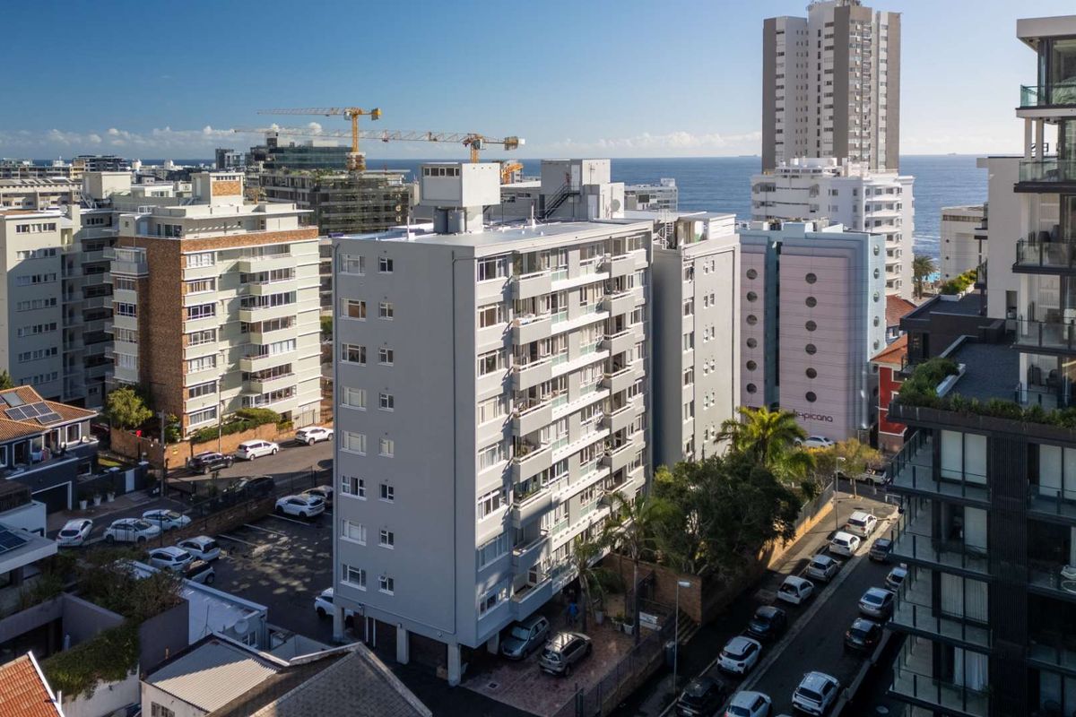 An elevated aerial view of a multi-story apartment building in a dense urban coastal setting, showcasing the building facade and surrounding neighborhood infrastructure.