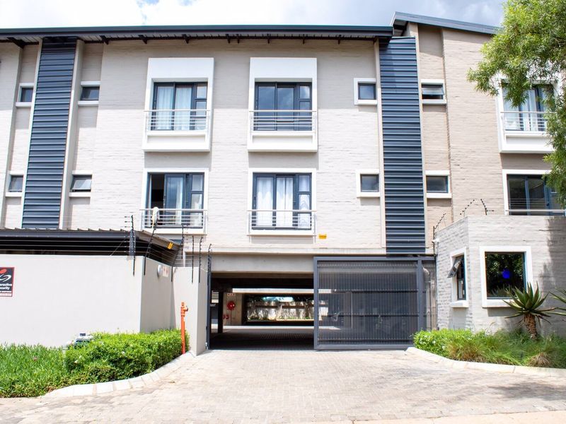 Front elevation of a modern apartment complex featuring a paved driveway, secure gated entrance, and balconies with metal cladding and electric fencing.