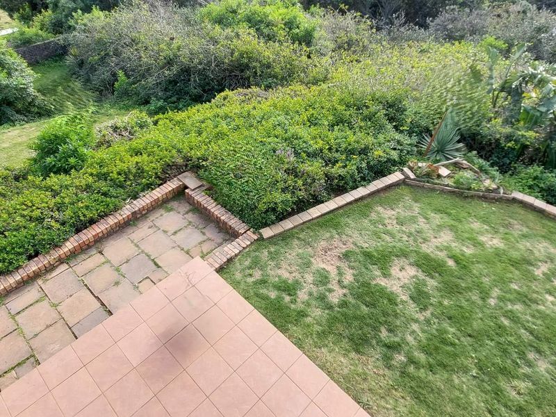An elevated view of the rear garden featuring a tiled patio area, brick retaining walls, and a lawn surrounded by established greenery.