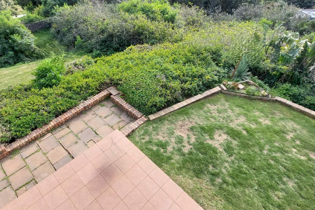 An elevated view of the rear garden featuring a tiled patio area, brick retaining walls, and a lawn surrounded by established greenery.