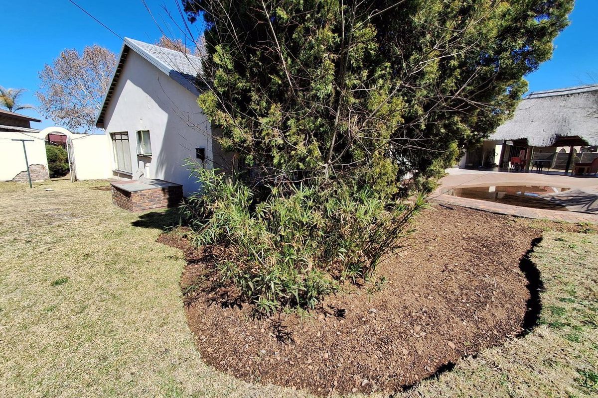 Rear garden area featuring a brick-built braai, landscaped garden beds, and a paved area leading to a thatched lapa and pool.