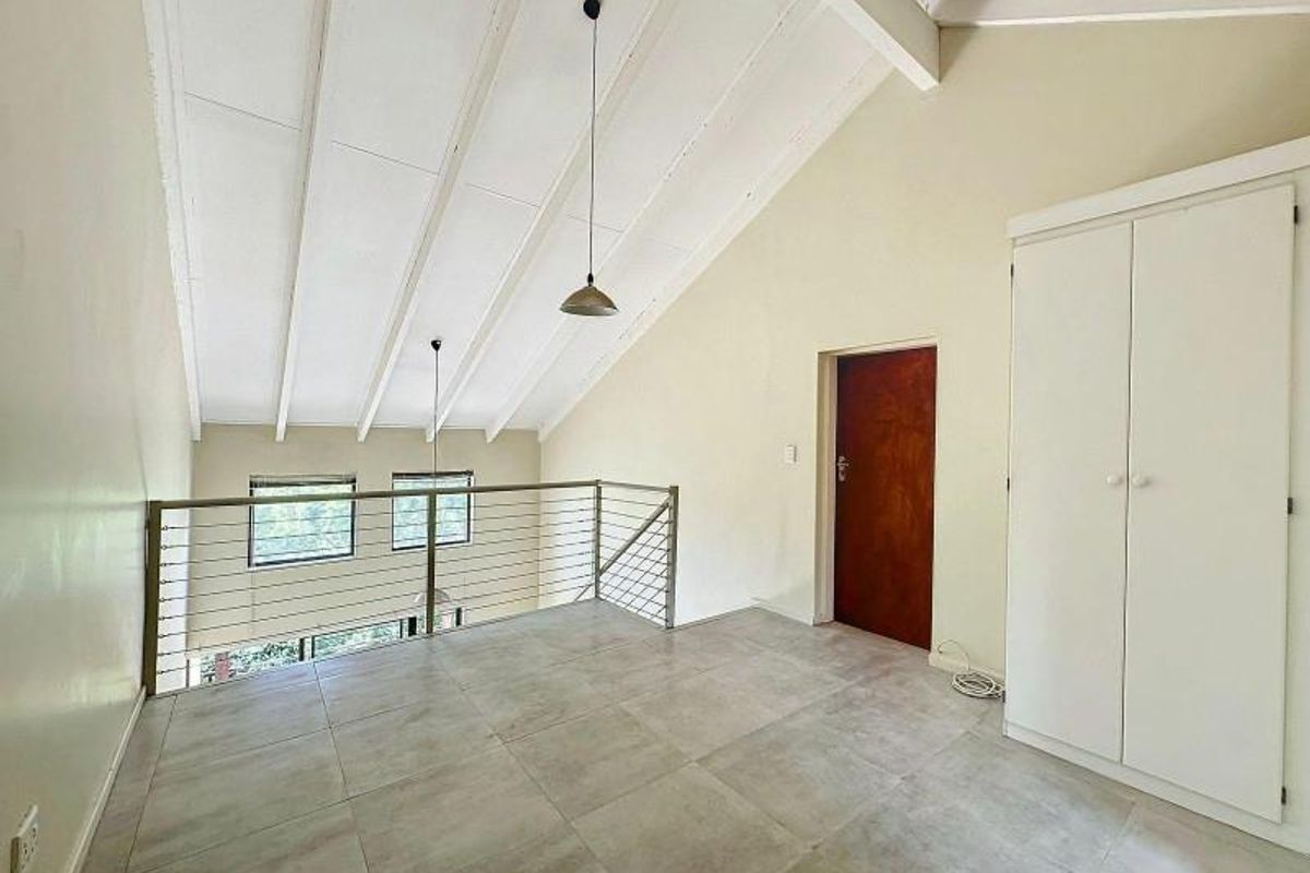 A spacious loft-style bedroom featuring vaulted ceilings with exposed rafters, tiled flooring, a built-in wardrobe, and a metal balustrade overlooking the lower level.