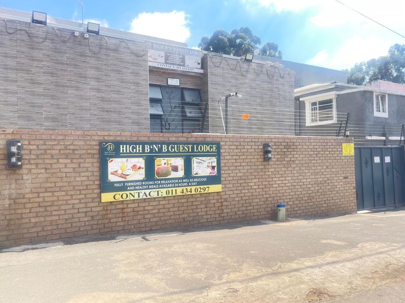 Front exterior view of the guest lodge featuring a brick boundary wall, electric fencing, and tiled facade cladding with prominent signage.