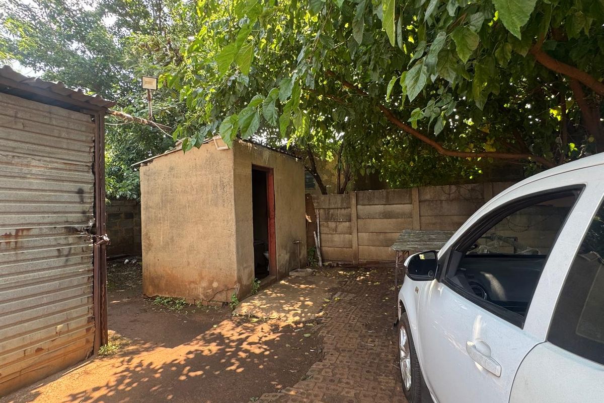 Rear exterior area featuring a small storage shed, precast concrete boundary walling, and paved driveway space under mature tree cover.