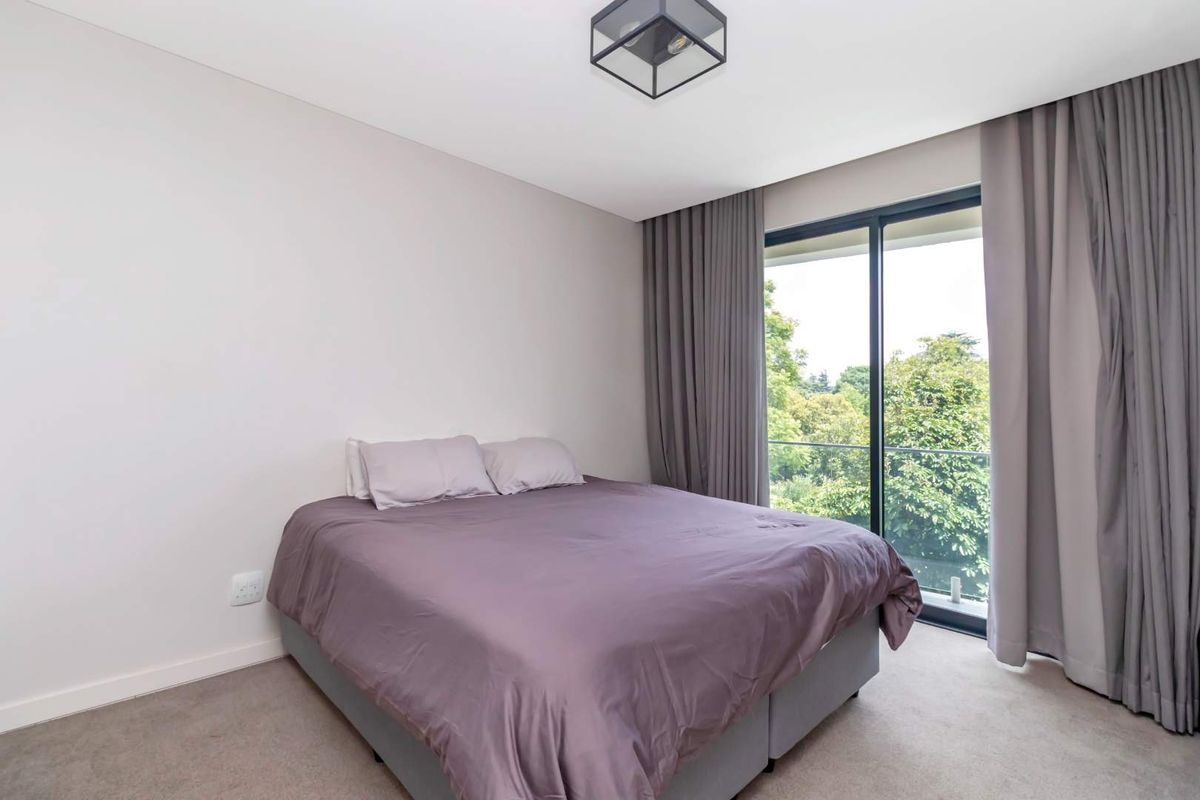 A well-maintained bedroom featuring neutral carpeting, a modern ceiling light, and large sliding glass doors opening onto a private balcony with leafy views.
