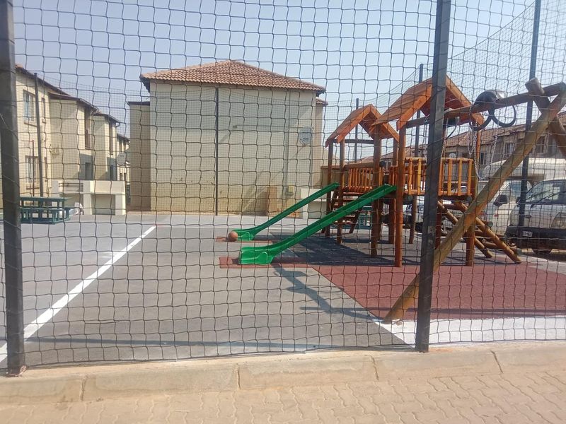 Communal outdoor area featuring a playground with slides and climbing structures, set within a fenced complex with paved surfaces.