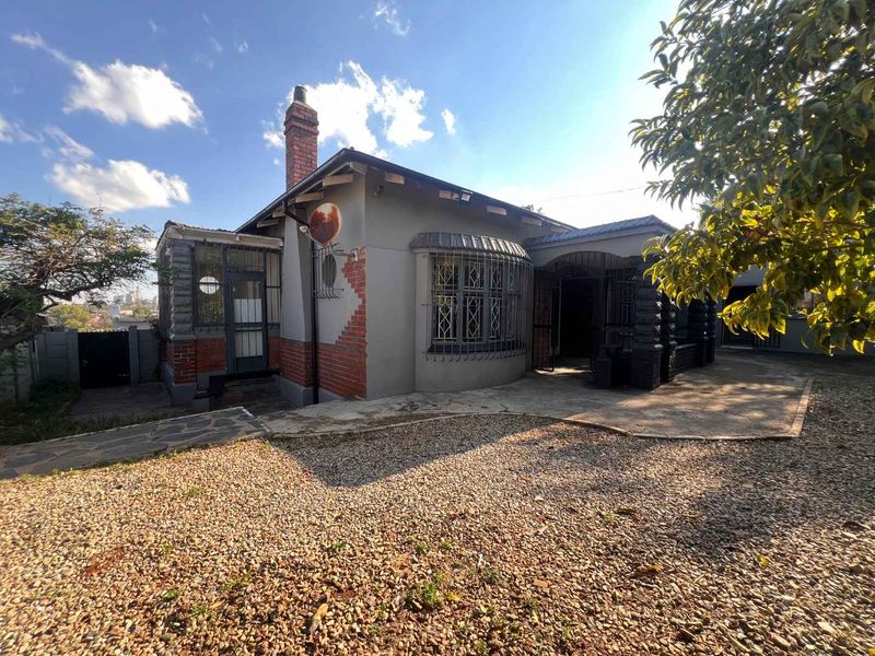 Front view of a single-story house featuring a brick chimney, security bars on windows, and a combination of paved and gravel driveway areas.