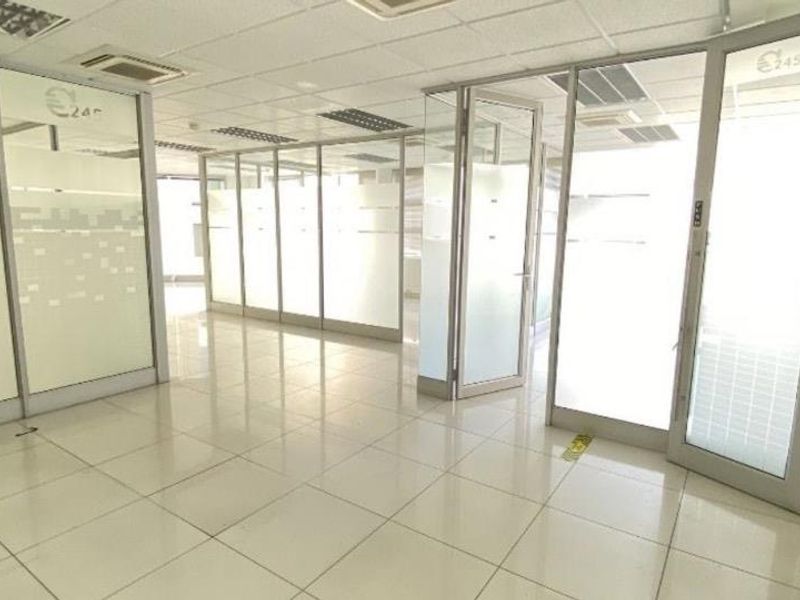 A bright, vacant office space featuring modern glass partitions, tiled flooring, and a suspended ceiling with integrated lighting.