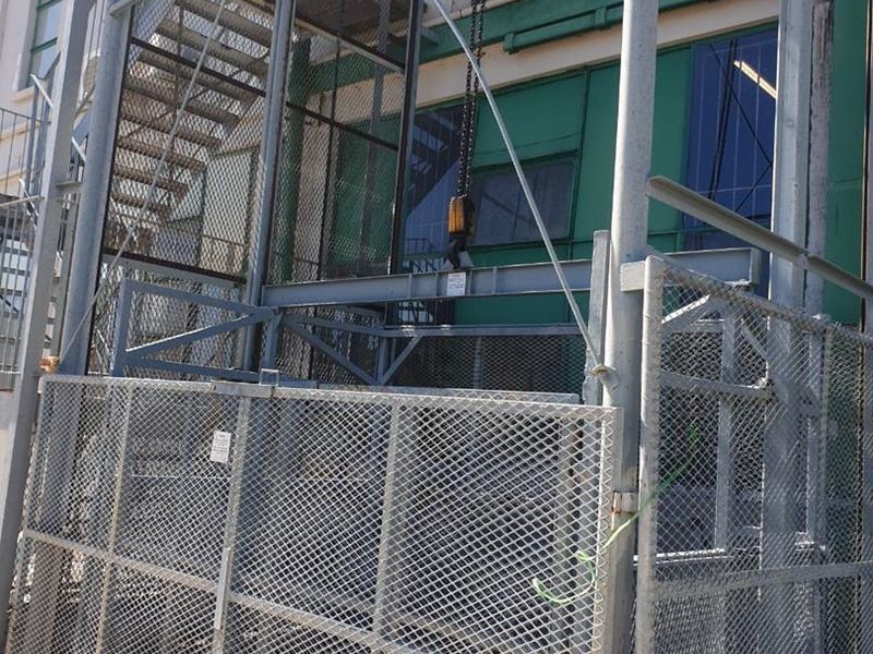Industrial exterior area featuring a metal goods lift, external staircase, and steel mesh security fencing against a painted industrial building facade.