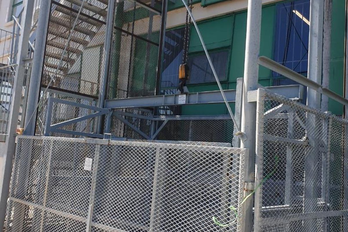 Industrial exterior area featuring a metal goods lift, external staircase, and steel mesh security fencing against a painted industrial building facade.