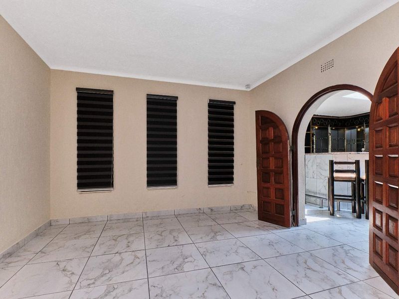 An empty living area featuring light-colored marble-look floor tiles, an arched wooden doorway, and modern roller blinds on the windows.