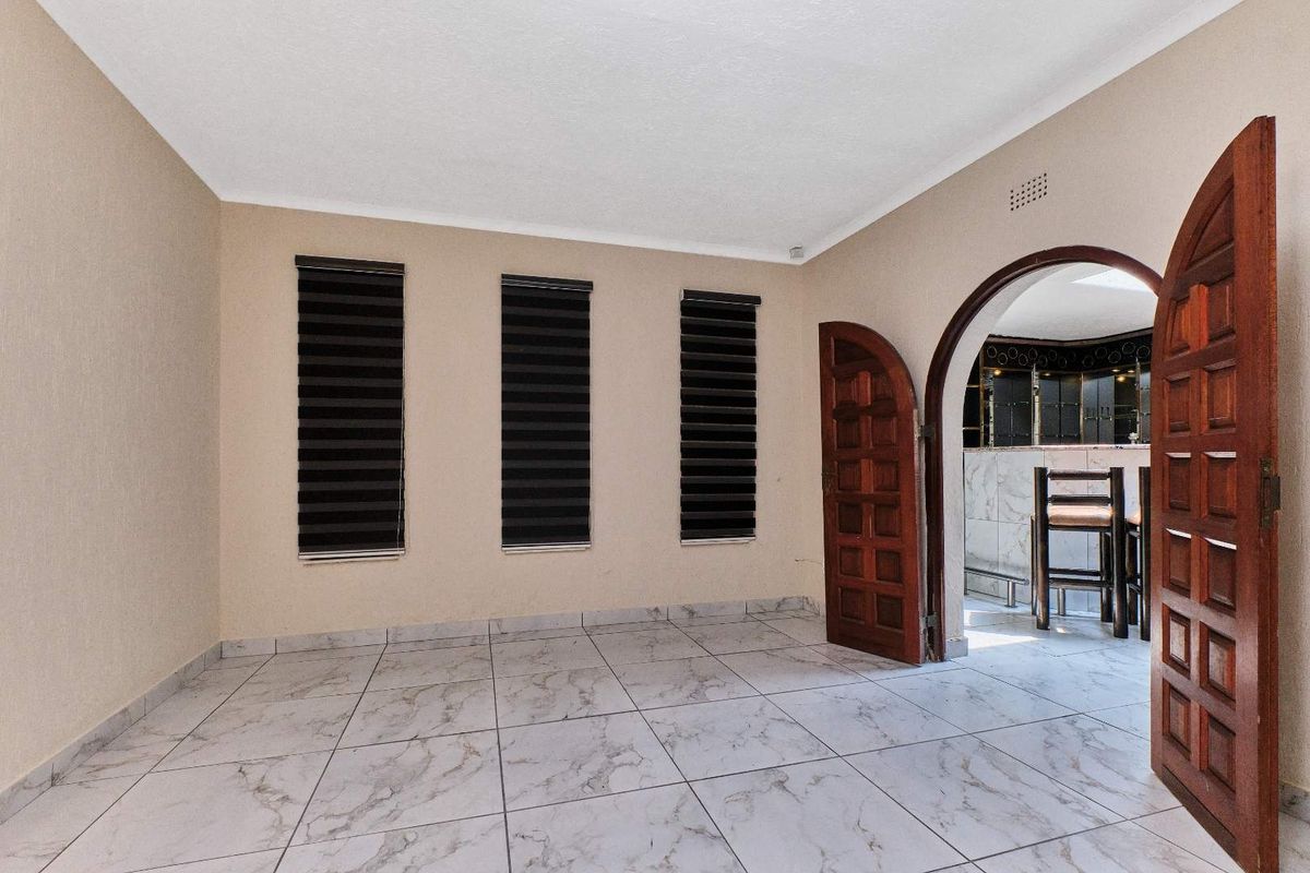 An empty living area featuring light-colored marble-look floor tiles, an arched wooden doorway, and modern roller blinds on the windows.