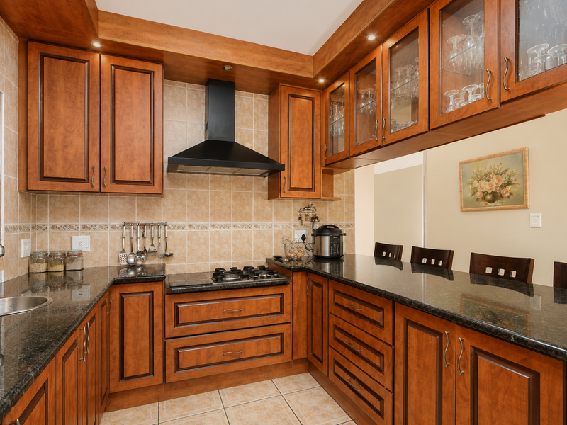 A well-maintained kitchen featuring warm-toned wooden cabinetry, granite countertops, a gas hob with an extractor fan, and integrated glass-fronted display cabinets.