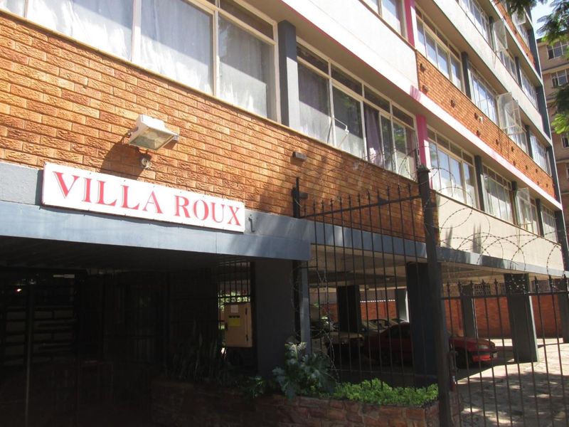 Front exterior view of Villa Roux apartment complex featuring brick facade, security fencing with razor wire, and covered parking access.