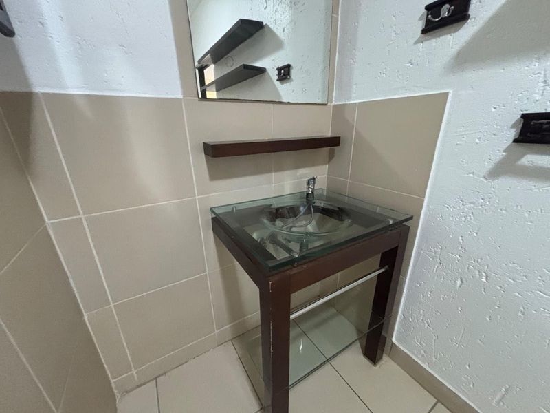 A compact bathroom featuring a modern glass vessel sink, vanity cabinet, wall mirror, and neutral wall and floor tiling.