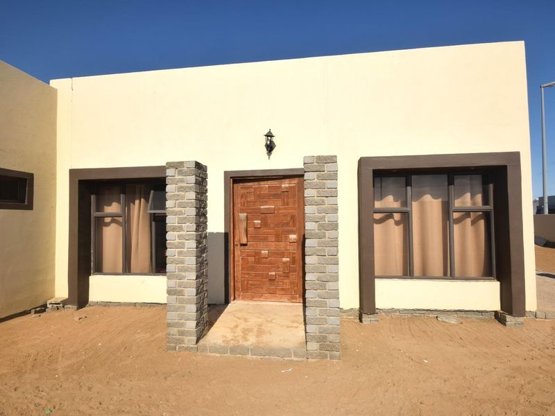 Front facade of a modern house featuring a timber entrance door, brick pillars, and dark aluminium window frames, set against an unfinished sandy yard.