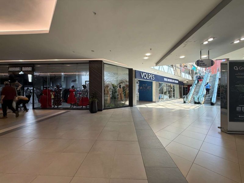 Interior view of a modern retail shopping mall corridor featuring tiled floors, glass storefronts, and an escalator leading to upper levels.