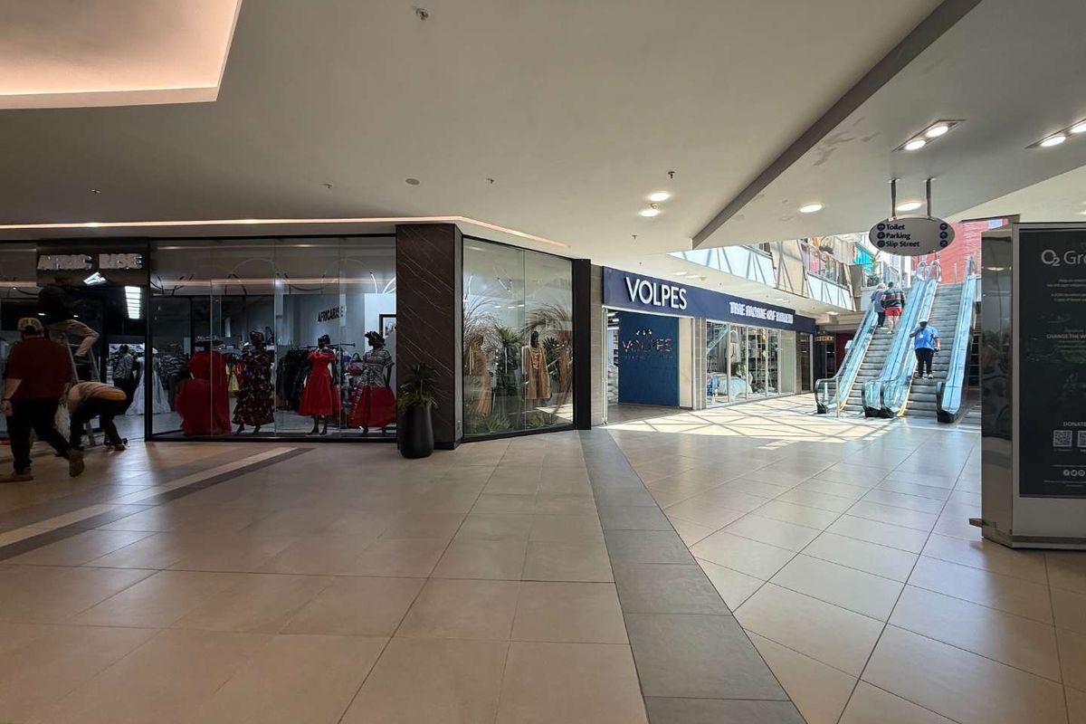 Interior view of a modern retail shopping mall corridor featuring tiled floors, glass storefronts, and an escalator leading to upper levels.
