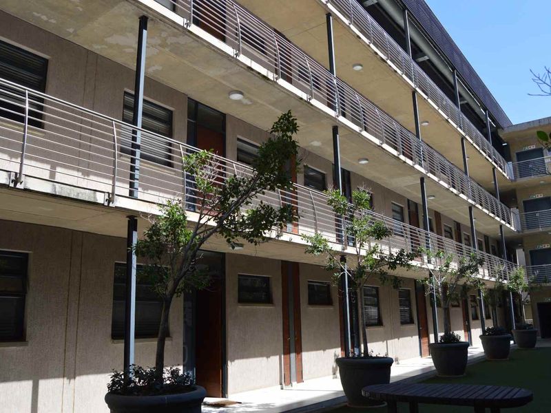 Exterior view of a multi-story apartment complex featuring concrete walkways, metal balcony railings, and a landscaped ground level area.