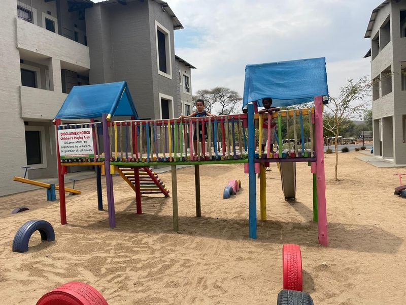 Communal outdoor playground area situated between apartment blocks, featuring fixed wooden play structures and a sand surface.