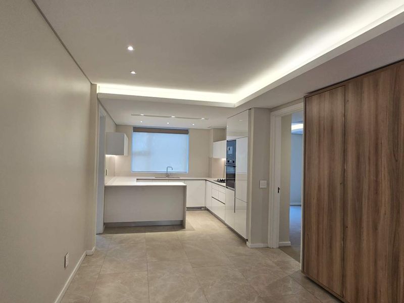 Modern, minimalist kitchen featuring sleek white cabinetry, stone countertops, integrated appliances, and sophisticated recessed cove lighting in an open-plan layout.