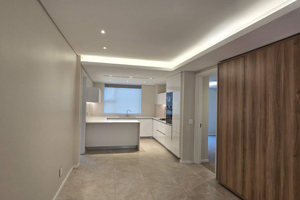 Modern, minimalist kitchen featuring sleek white cabinetry, stone countertops, integrated appliances, and sophisticated recessed cove lighting in an open-plan layout.