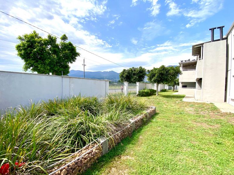 Rear exterior view of the apartment complex featuring a landscaped garden with a gabion retaining wall, perimeter fencing, and mountain views.