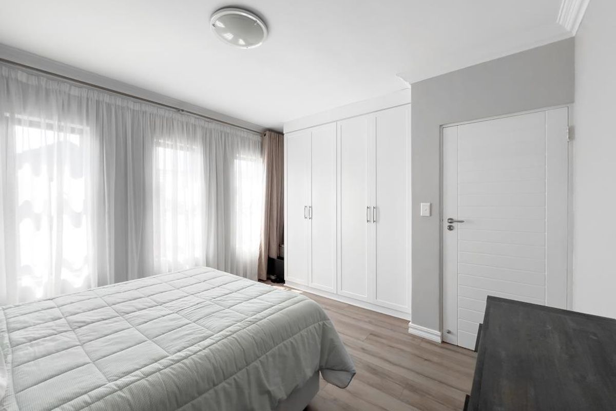 A bright and well-maintained bedroom featuring built-in wardrobes, modern laminate flooring, and large windows with sheer curtains.