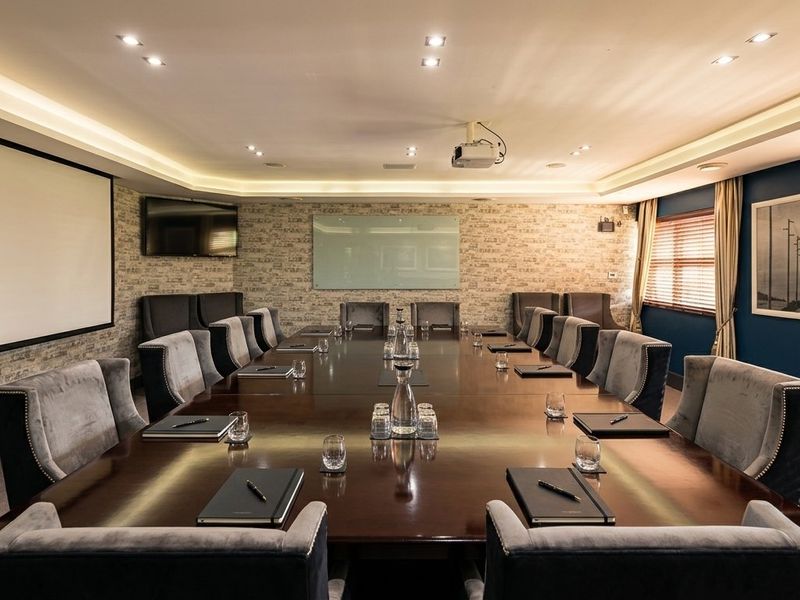 A professional boardroom featuring a large conference table, integrated projector system, wall-mounted screen, whiteboard, and sophisticated recessed and cove lighting.